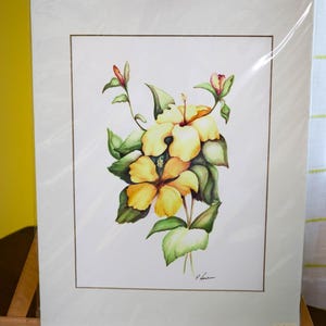 May include: Watercolor painting of yellow hibiscus flowers with green leaves and red buds. The artwork is framed and displayed on a wooden easel. The painting is signed by the artist.