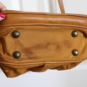 Pu&ograve; includere: Primo piano del fondo di una borsa a mano marrone con quattro piedini in metallo color ottone. La borsa ha un bordo in pelle marrone e un corpo in tela marrone. Ci sono macchie scure sulla tela.