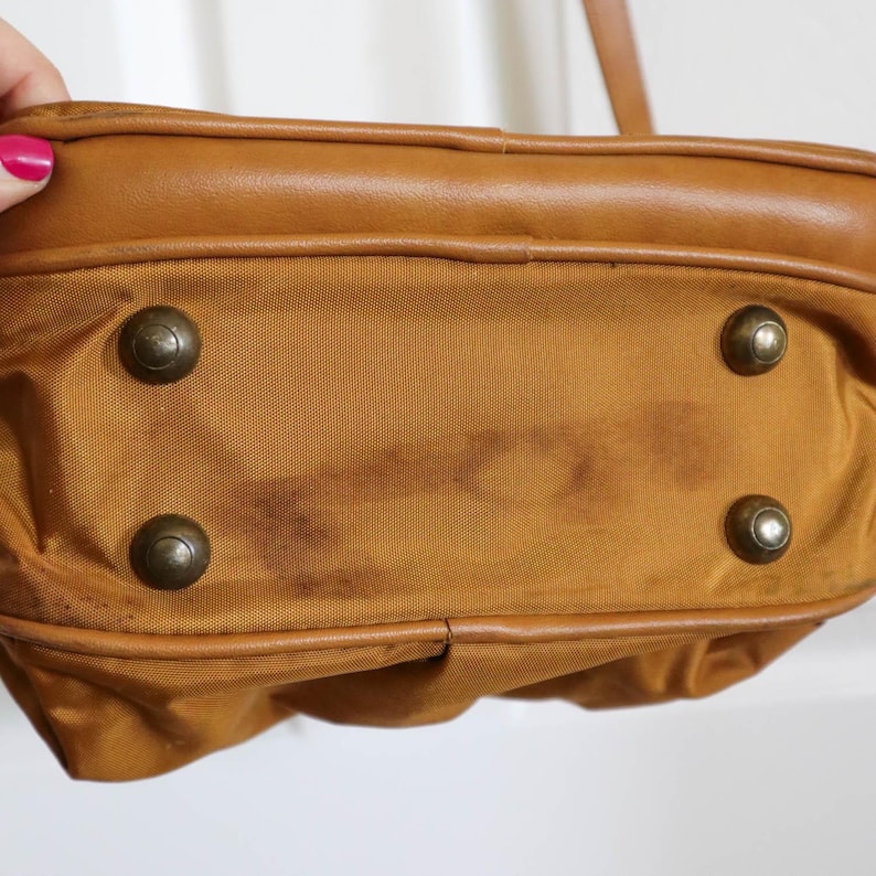 Pu&ograve; includere: Primo piano del fondo di una borsa a mano marrone con quattro piedini in metallo color ottone. La borsa ha un bordo in pelle marrone e un corpo in tela marrone. Ci sono macchie scure sulla tela.