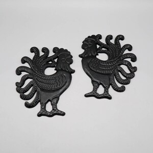 May include: Two black cast iron rooster silhouettes featuring detailed feather patterns on the tails and wings. Each rooster stands in profile with a textured surface, showcasing a distinct comb and wattle design, presented as a pair of matching items.