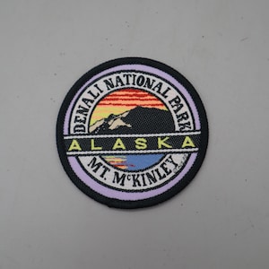 May include: A circular embroidered patch featuring a landscape of Denali National Park in Alaska. The design includes a mountain silhouette against a sunset sky, with the text Denali National Park, Alaska, and Mt. McKinley in black lettering.