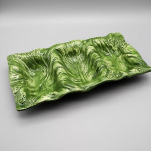 May include: A green ceramic serving dish with a wavy, leaf-like design. The rectangular dish has a glossy finish and a textured surface, with a light green and white color variation. The dish is designed to resemble a large leaf.