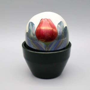 Op de afbeelding: Een decoratief keramisch ei met een rood tulpontwerp, rustend in een donkergroene keramische houder. Het ei heeft een witte achtergrond met blauwe en groene bloemaccenten. De houder heeft een eenvoudige, afgeronde vorm.