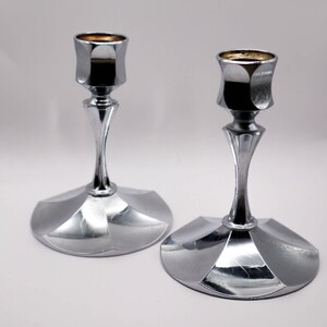 May include: Two polished silver candle holders. Each has a faceted cup, a slender stem, and a geometric base. The candle cups have a dark interior. The candle holders are displayed on a white surface.
