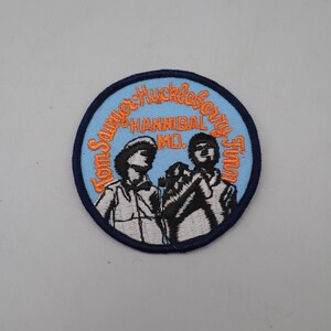 May include: A circular embroidered patch with a light blue background and a dark blue border. It features black and white illustrations of two figures with the orange embroidered text Tom Sawyer-Huckleberry Finn and HANNIBAL MO.