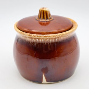 May include: A brown ceramic jar with a lid. The jar has a rounded shape with a glossy finish. The lid has a knob on top. The rim of the jar and the lid's edge have a tan color. The jar is likely for kitchen use.