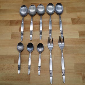 May include: A collection of stainless steel silverware, including spoons and forks, arranged on a wooden surface. The silverware features a decorative design on the handles. The spoons are of varying sizes, and the forks have four tines.