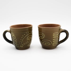 May include: Two ceramic mugs with a dark olive-green exterior and brown interior. Each mug features a decorative design in a lighter olive-green color. The designs include spirals, dots, and curved lines. The mugs have black handles.