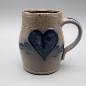 Può includere: Tazza in gres con un grande cuore blu. La tazza ha un manico e una forma leggermente svasata. Il cuore è dipinto in stile folk, con alcuni accenti di foglie blu. La tazza è di colore beige neutro.