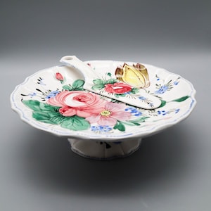 Puede incluir: Un soporte para pasteles de cerámica blanca con un diseño floral con rosas rosas, hojas verdes y detalles azules. Un servidor a juego descansa sobre el soporte. El soporte tiene un borde festoneado y una base de pedestal.