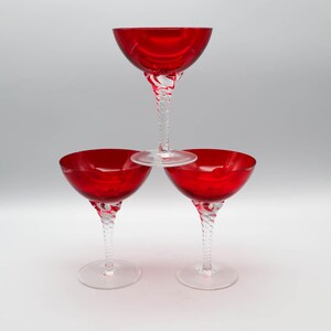 Puede incluir: Tres copas tipo coupe con cuencos de color rojo intenso y tallos de vidrio transparente retorcido. Las copas están dispuestas en formación triangular, con una sobre las otras dos, todas con una base circular lisa y un borde ancho.