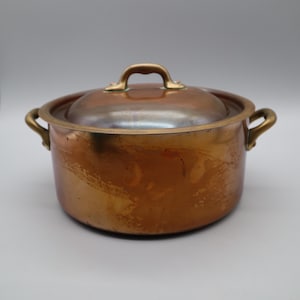 May include: A round copper pot with a matching lid and two side handles. The exterior surface of the pot and lid shows a warm, metallic copper finish with visible signs of use, including subtle surface variations and a polished brass handle.