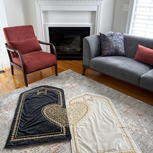 May include: Two prayer rugs, one black and one cream, with gold embroidered detailing. The rugs feature a heart-shaped design and a decorative border. The rugs are placed on a patterned rug in a living room setting.
