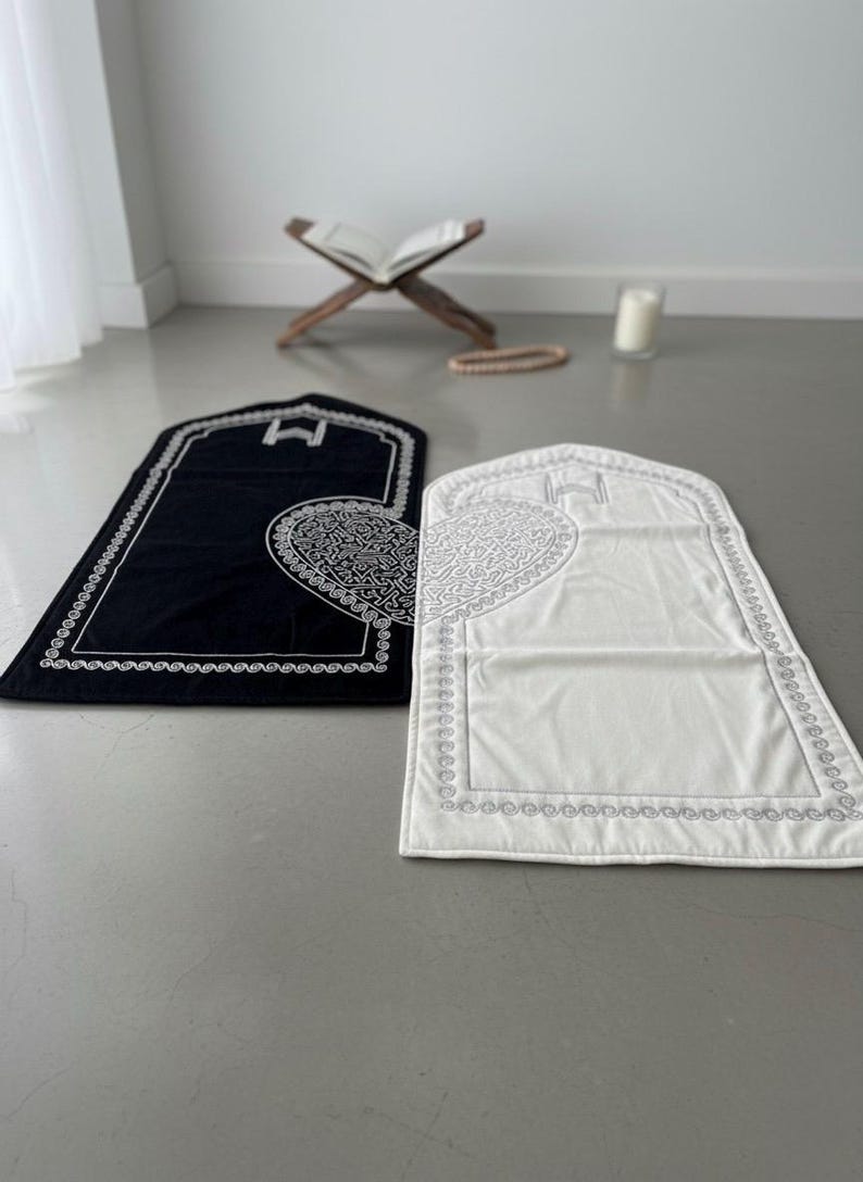 May include: Two prayer mats, one black and one white, are displayed on a gray floor. Each mat has a decorative border and a heart-shaped design in the center. A wooden book stand and a string of prayer beads are in the background.