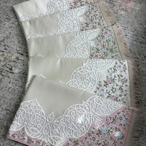 K&ouml;nnte beinhalten: Mehrere Gebetsteppiche mit aufwendigen wei&szlig;en Stickereien und floralen Bord&uuml;ren in Rosa, Blau und Gr&uuml;n. Die Teppiche haben einen cremefarbenen Hintergrund und gefranste Kanten. Die Teppiche sind f&auml;cherf&ouml;rmig angeordnet.