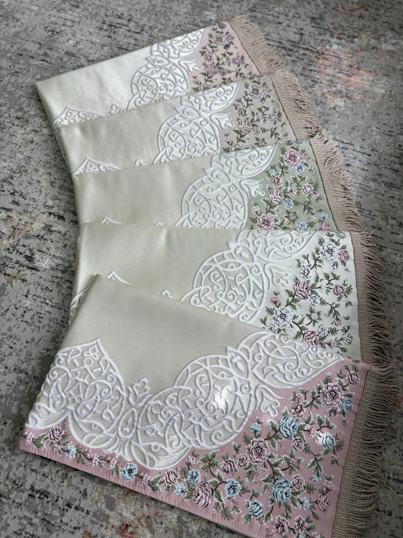 K&ouml;nnte beinhalten: Mehrere Gebetsteppiche mit aufwendigen wei&szlig;en Stickereien und floralen Bord&uuml;ren in Rosa, Blau und Gr&uuml;n. Die Teppiche haben einen cremefarbenen Hintergrund und gefranste Kanten. Die Teppiche sind f&auml;cherf&ouml;rmig angeordnet.