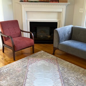 K&ouml;nnte beinhalten: Ein Gebetsteppich mit hellgrauer Mitte und einem rosa Rand mit floralen und verzierten Golddesigns. Der Teppich liegt auf einem gemusterten Teppich, mit einem roten Sessel und einem grauen Sofa im Hintergrund.