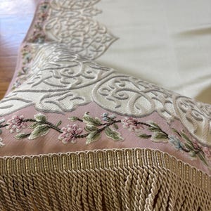 K&ouml;nnte beinhalten: Nahaufnahme eines dekorativen Textils mit aufwendiger Stickerei. Das Design zeigt Blumen- und Scrollmuster in Creme, Rosa und Gr&uuml;n. Der Rand ist mit einer beigen Franse versehen.