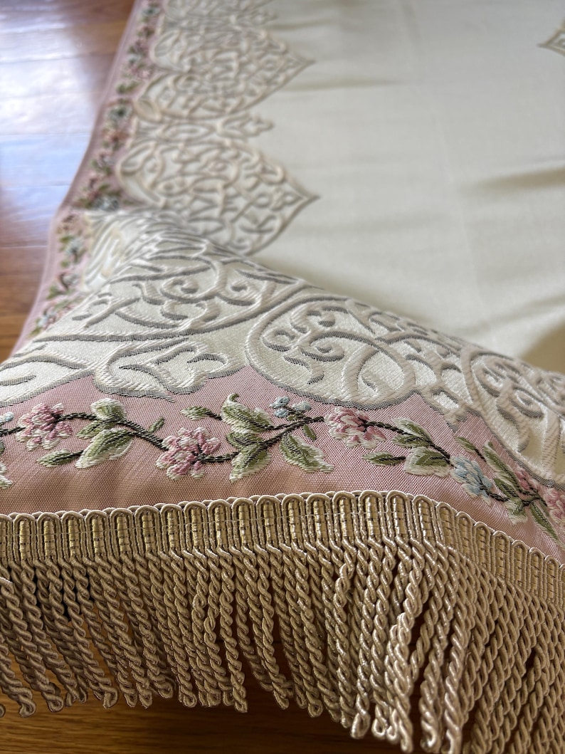 K&ouml;nnte beinhalten: Nahaufnahme eines dekorativen Textils mit aufwendiger Stickerei. Das Design zeigt Blumen- und Scrollmuster in Creme, Rosa und Gr&uuml;n. Der Rand ist mit einer beigen Franse versehen.