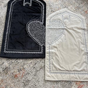 May include: Two prayer mats, one black and one cream, are displayed. Each mat features a decorative border and a heart-shaped design with intricate patterns. The mats have a rectangular shape with an arched top, and are likely used for religious practices.