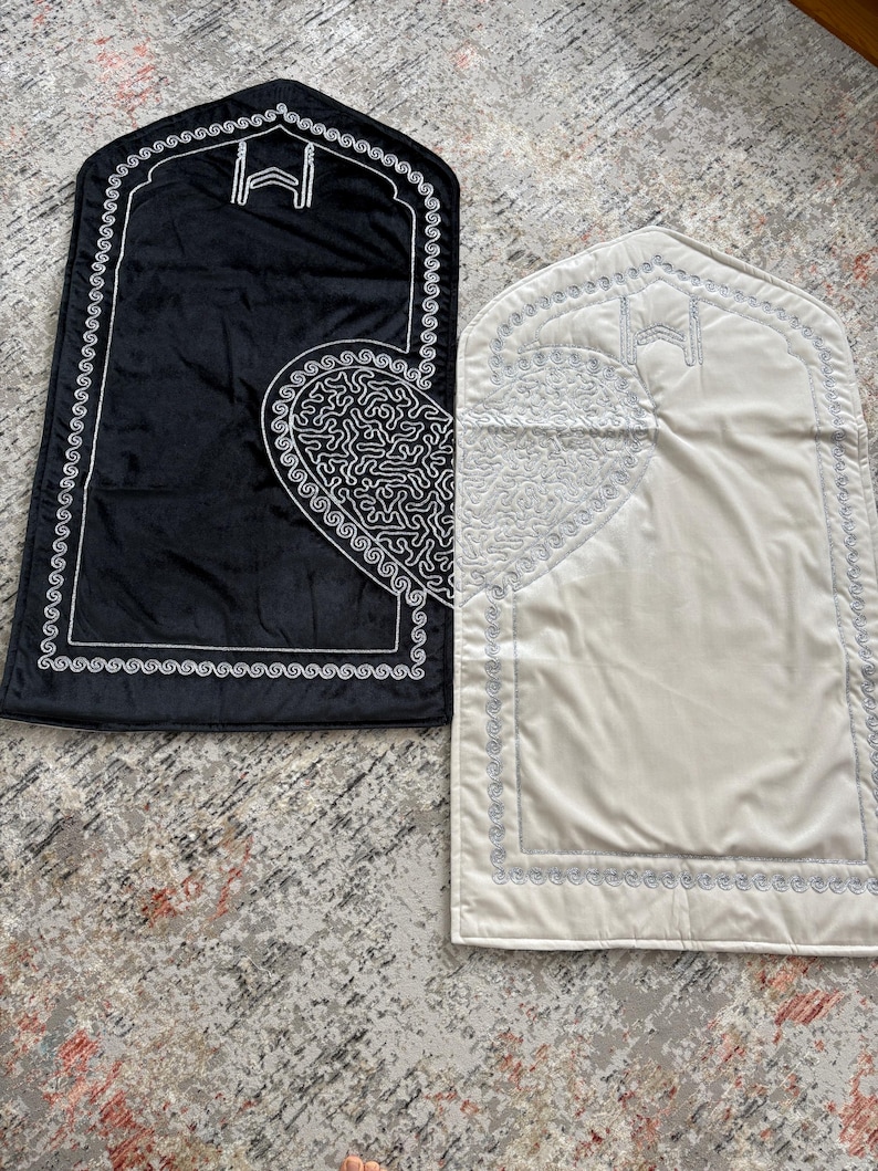 May include: Two prayer mats, one black and one cream, are displayed. Each mat features a decorative border and a heart-shaped design with intricate patterns. The mats have a rectangular shape with an arched top, and are likely used for religious practices.