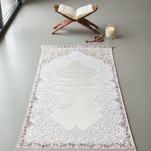 K&ouml;nnte beinhalten: Gebetsteppich mit einem wei&szlig;en Mittelfeld und einem dekorativen Rand in Rosa- und Lilat&ouml;nen. Der Teppich weist ein verziertes, gepr&auml;gtes Design auf. Ein h&ouml;lzerner Buchst&auml;nder, eine brennende Kerze und Gebetsketten befinden sich im Hintergrund.