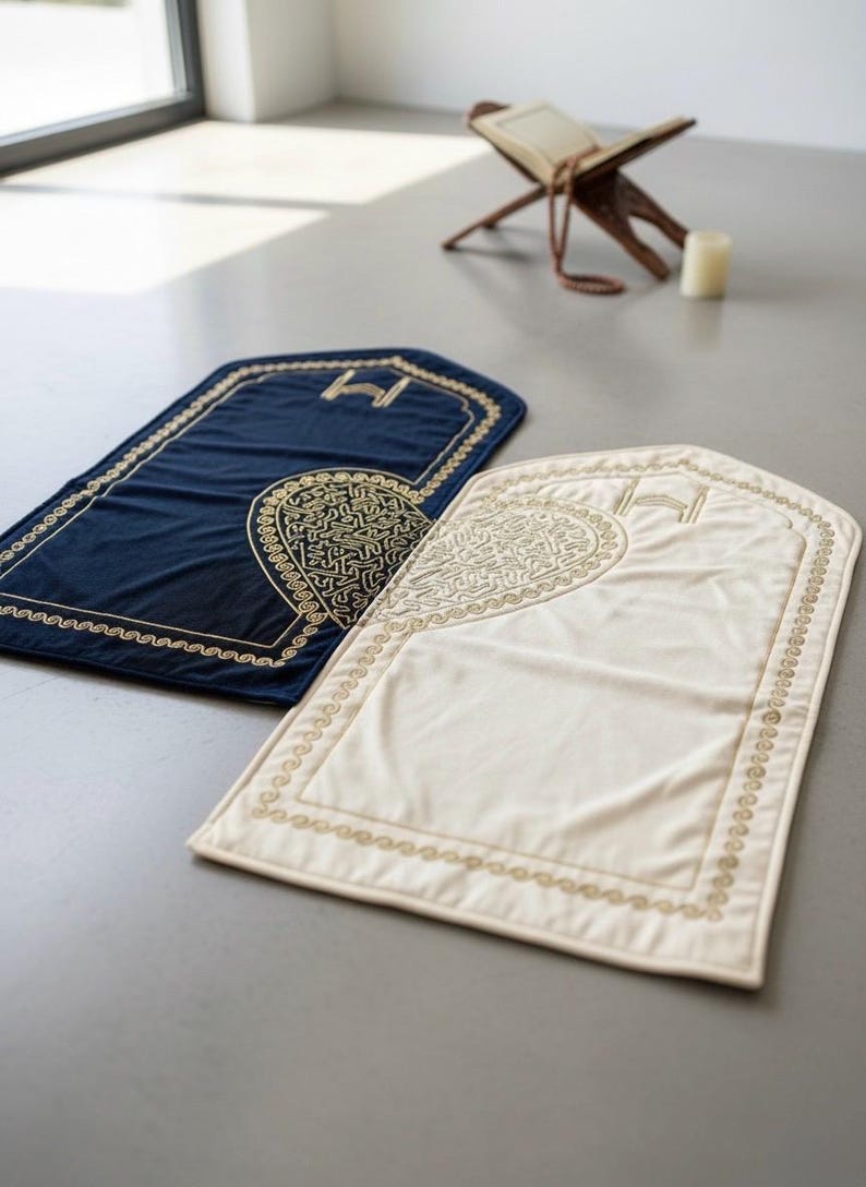 May include: Two prayer rugs, one navy blue and one cream, are displayed on a gray surface. Both rugs have a rectangular shape with an arched top and are adorned with gold embroidery. A wooden book stand and candle are in the background.