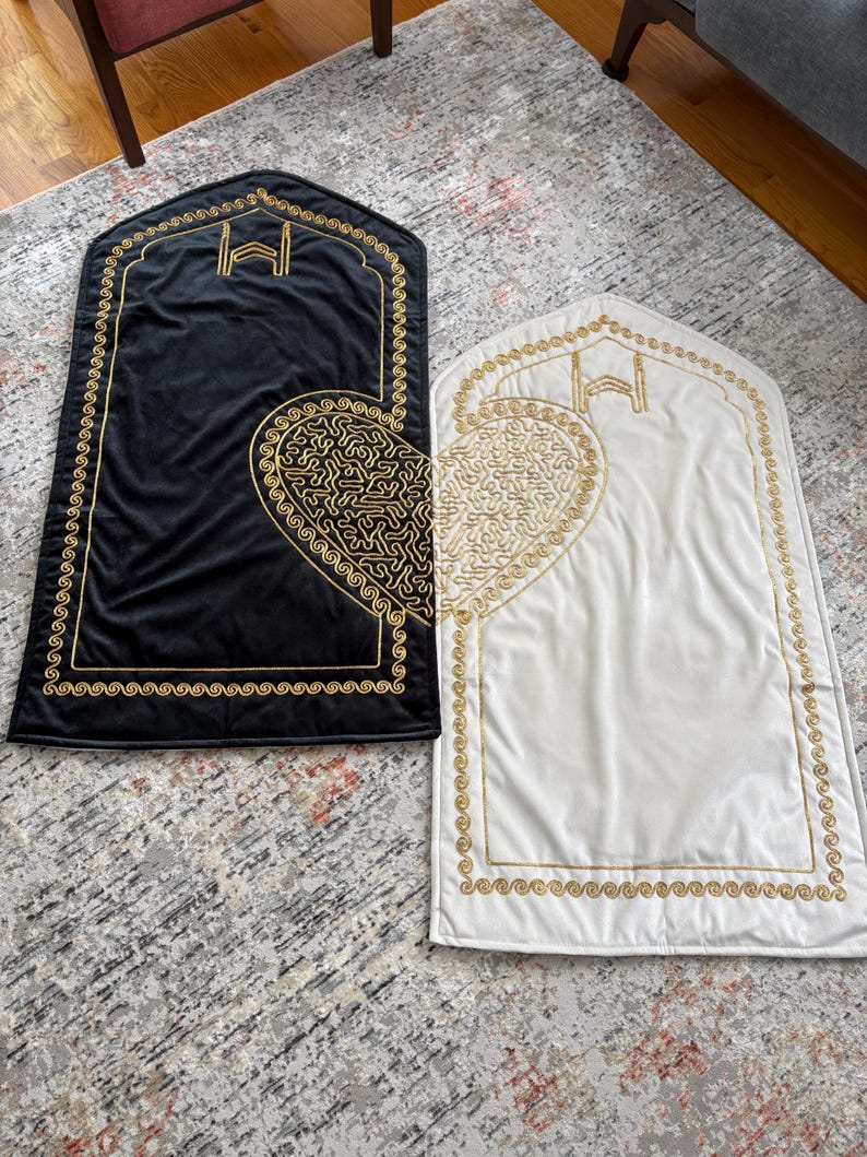 May include: Two prayer rugs, one black and one white, are displayed side-by-side. Each rug features a gold embroidered heart design and a gold border. The top of each rug has a gold embroidered arch design.