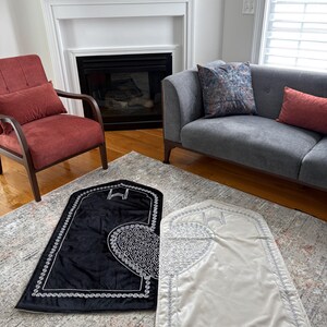 May include: Two prayer rugs, one black and one cream, are displayed on a patterned rug. The black rug features a white, ornate design and border. The cream rug has a similar design with a silver border. The rugs are rectangular with an arched top.