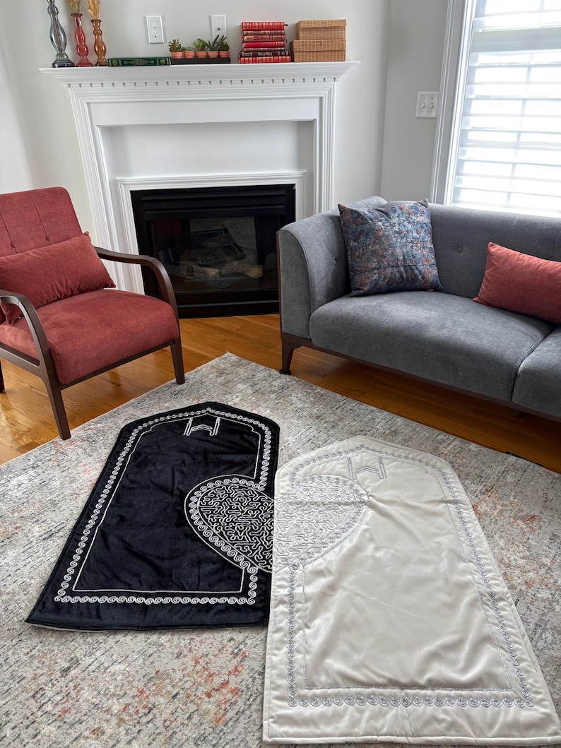 May include: Two prayer rugs, one black and one cream, are displayed on a patterned rug. The black rug features a white, ornate design and border. The cream rug has a similar design with a silver border. The rugs are rectangular with an arched top.