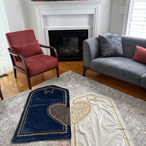 May include: Two prayer rugs, one navy blue and one cream, are displayed on a patterned rug. Both rugs have a gold border and a gold heart-shaped design. The background includes a living room with a fireplace, chair, and sofa.