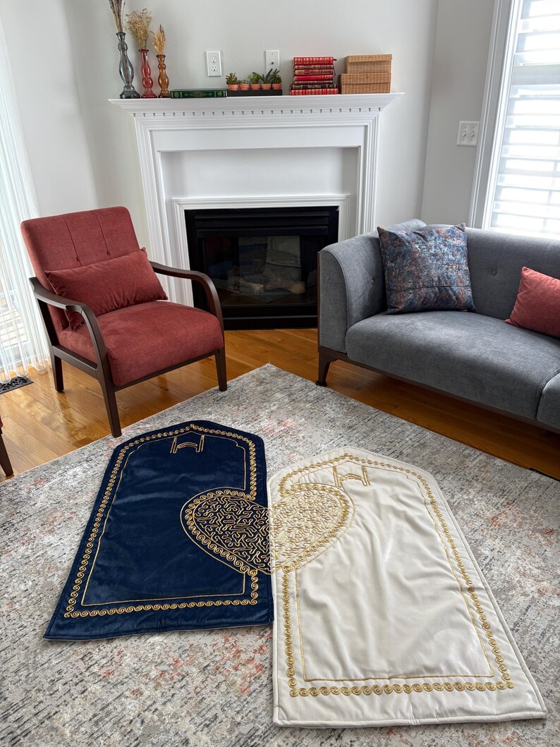 May include: Two prayer rugs, one navy blue and one cream, are displayed on a patterned rug. Both rugs have a gold border and a gold heart-shaped design. The background includes a living room with a fireplace, chair, and sofa.