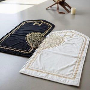 May include: Two prayer mats, one black and one white, are displayed. Each mat has a gold embroidered border and a central design. A wooden book stand and a candle are in the background.
