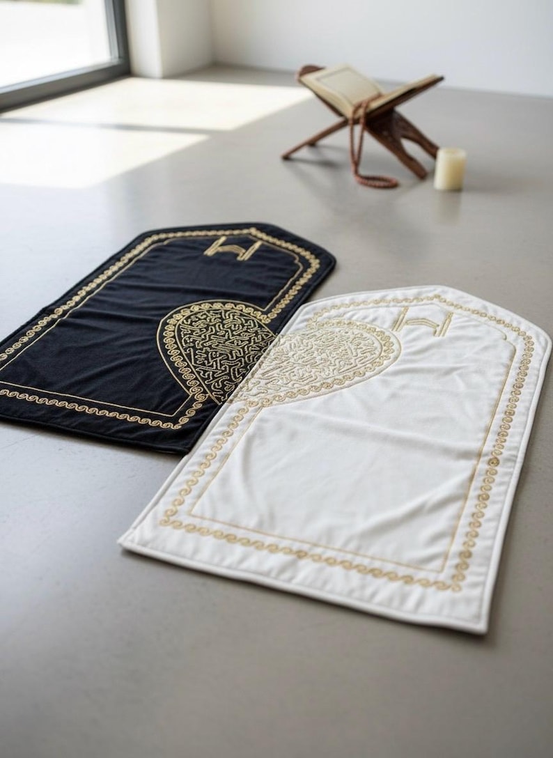 May include: Two prayer mats, one black and one white, are displayed. Each mat has a gold embroidered border and a central design. A wooden book stand and a candle are in the background.