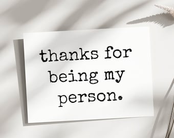 May include: A white card with the text "thanks for being my person" in a typewriter-style font. The card is on a white surface with shadows and a sprig of dried plant material.