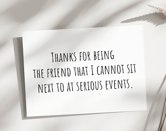May include: A white card with black text that reads "THANKS FOR BEING THE FRIEND THAT I CANNOT SIT NEXT TO AT SERIOUS EVENTS."