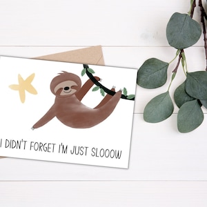 May include: A white card with a brown envelope behind it. The card features a cartoon sloth hanging from a branch with a yellow star in the background. The text on the card reads "I didn't forget I'm just sloooow".