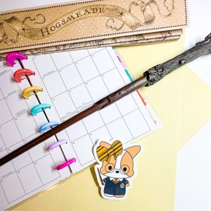 May include: A brown wooden wand with a black handle, a yellow and brown sticker of a cartoon dog wearing a Hogwarts uniform and a yellow bow, and a calendar with colorful tabs. The calendar has the word "April" written on it.