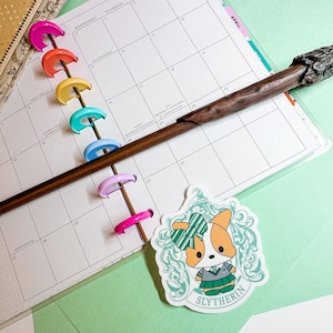 May include: A sticker featuring a cartoon corgi wearing a green and gold Slytherin house uniform. The corgi is surrounded by a green and gold crest with the words "Slytherin" printed below.