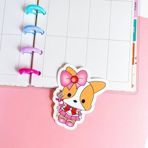 Könnte beinhalten: Ein Aufkleber mit einem Cartoon-Corgi, der ein rosa-weißes Matrosen-Outfit trägt und einen rosa-goldenen Zauberstab hält. Der Corgi hat eine rosa Schleife auf dem Kopf und ein weißes Gesicht mit schwarzen Augen.