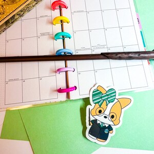 May include: A sticker of a cartoon dog wearing a black and green Hogwarts uniform with a green and white bow tie. The dog is holding a wand in its right paw. The sticker is on a green background.