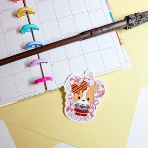 May include: A sticker featuring a cartoon corgi wearing a Gryffindor uniform and a red and gold bow. The sticker is on a yellow background with a calendar and a wand in the background.