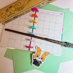 May include: A 2022 planner with colorful binding rings, a wooden wand, and a sticker of a corgi wearing glasses and a wizard robe. A box with the text "HOGSMEADE" is in the background. Green paper is also visible.