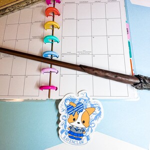 May include: A colorful monthly calendar with a wooden wand laying across the pages. The calendar has a sticker of a cartoon corgi wearing a blue and white uniform with the text "RAVENCLAW" below it.