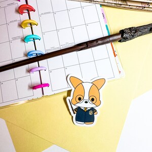 May include: A sticker of a cartoon corgi wearing glasses and a dark blue robe, holding a wand. A wooden wand and a planner with colorful rings are also visible. The background is yellow and white.