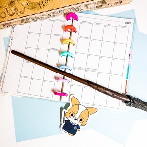 May include: A monthly planner with colorful binder clips and a brown wooden wand lying on top. The planner has a blue background and the month of March is visible. A sticker of a cartoon corgi wearing a blue uniform is on the bottom right corner.