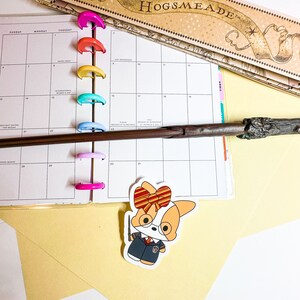 May include: A planner with colorful binder rings, a wand, and a sticker of a corgi wearing a school uniform and holding a wand. A map of Hogsmeade is visible in the background. The planner is open to the month of April.