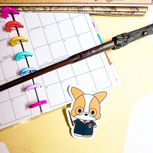 May include: A white and orange cartoon corgi wearing a blue and gold Hogwarts uniform and holding a brown and black wand. The corgi is on a white sticker with a white background.