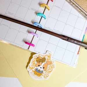 May include: A yellow and gold sticker with a cartoon corgi wearing a yellow and black bow, a yellow and black uniform, and a yellow and black crest. The crest says "Hufflepuff".