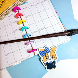 May include: A white sticker with a cartoon dog wearing a blue and white striped bow and a blue and black uniform. The dog is holding a brown and black wand. The sticker is placed on a white planner with colorful rings.